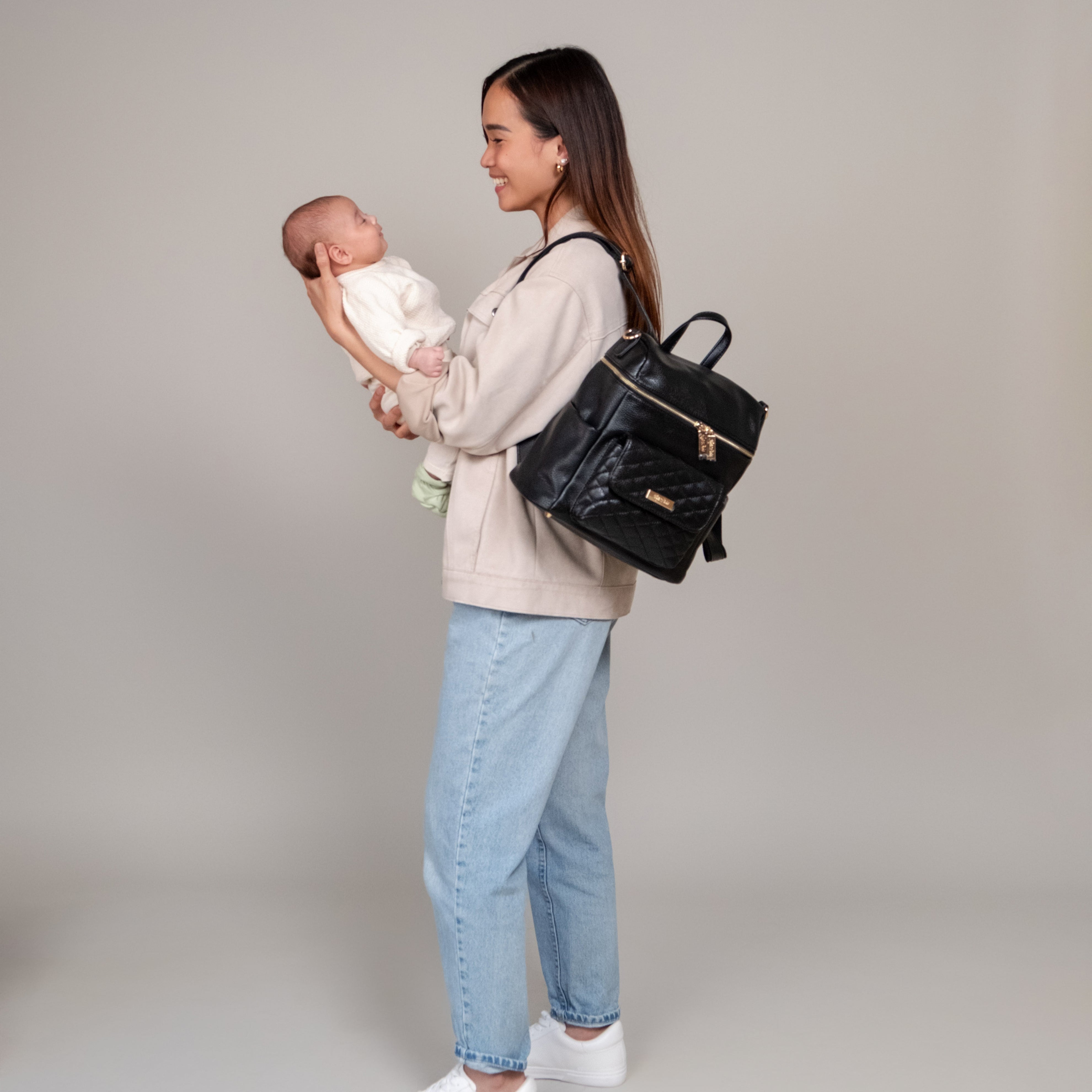 Mom holding baby and wearing Petit Monaco ebony black diaper bag by Luli Bebe on shoulder, stylish compact diaper backpack for baby essentials