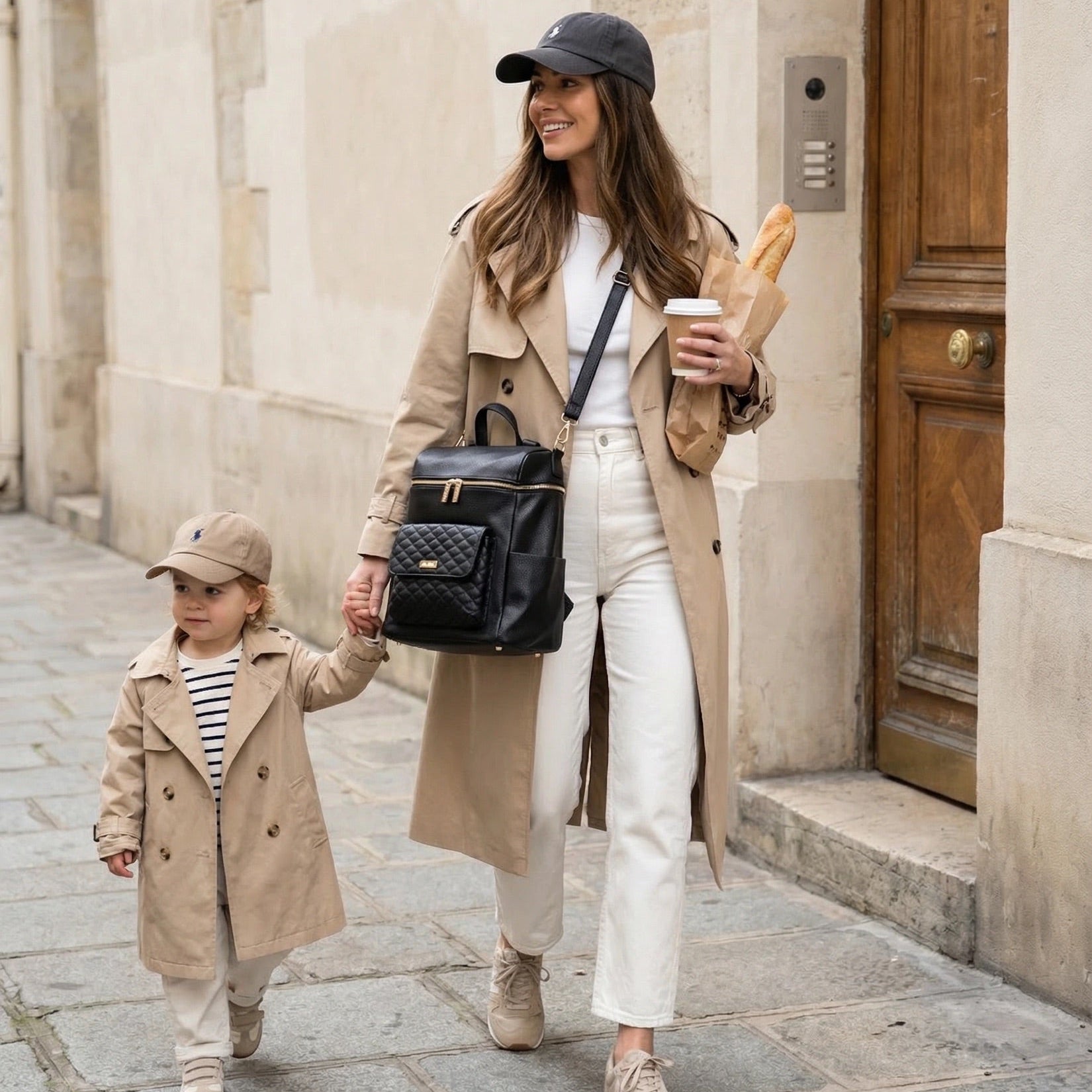 Woman and child walking together on a street, both wearing beige coats and hats, with the woman holding a black diaper bag backpack and a coffee cup.